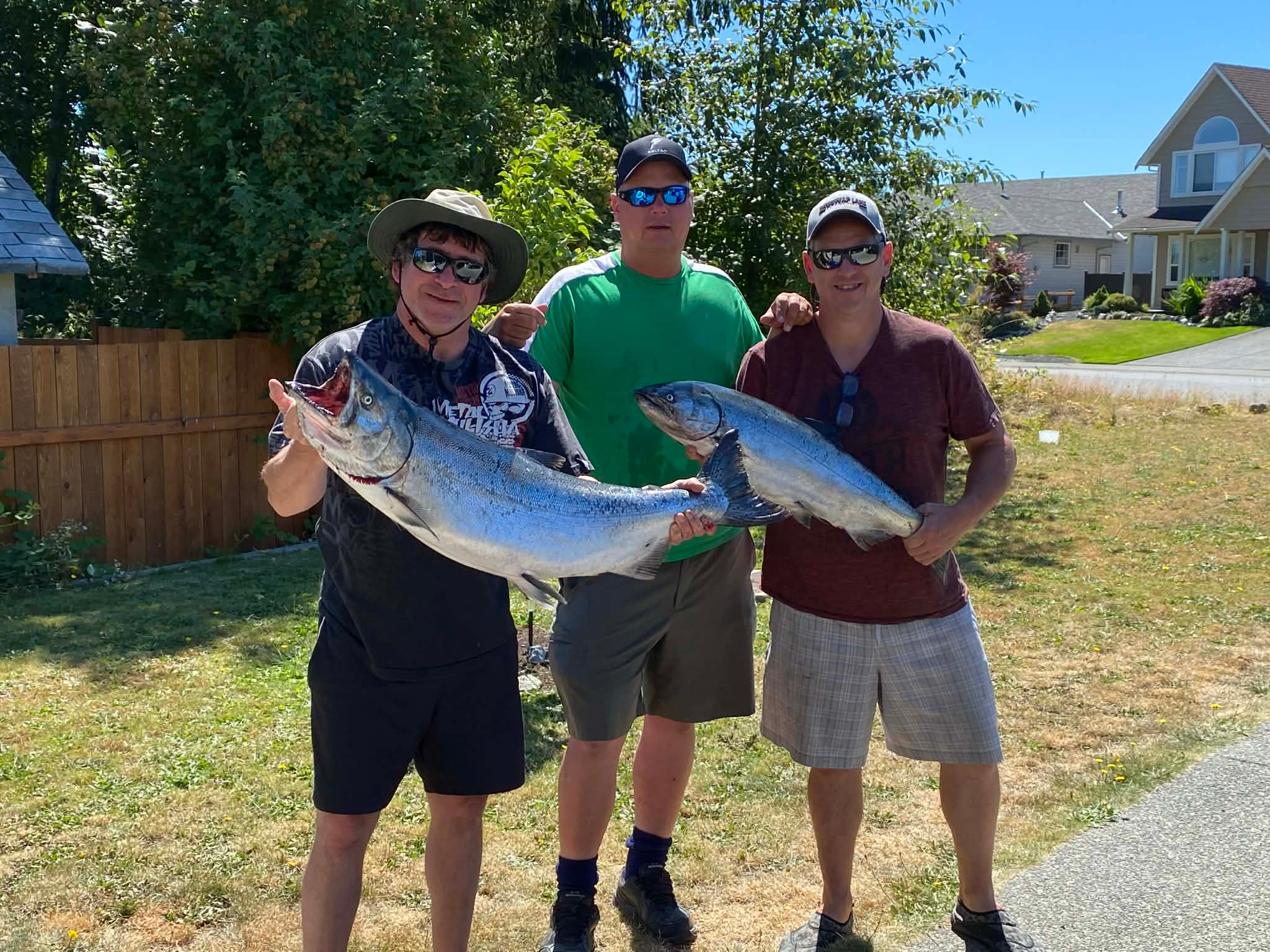 Three men pose on a lawn holding a pair of large, silver-colored fish. They are smiling and houses, fences and trees are visible in the background.